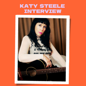 Photo of Katy Steele looking into camera with arm resting on black guitar against an orange background.